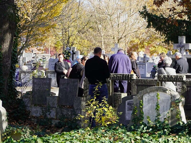 Parast compared cu peisajul vibrant din oraș, imaginea arată o adunare de oameni în cimitirul din Sibiu, în timpul toamnei, într-o zi însorită, cu frunziș colorat în nuanțe de auriu și portocaliu.